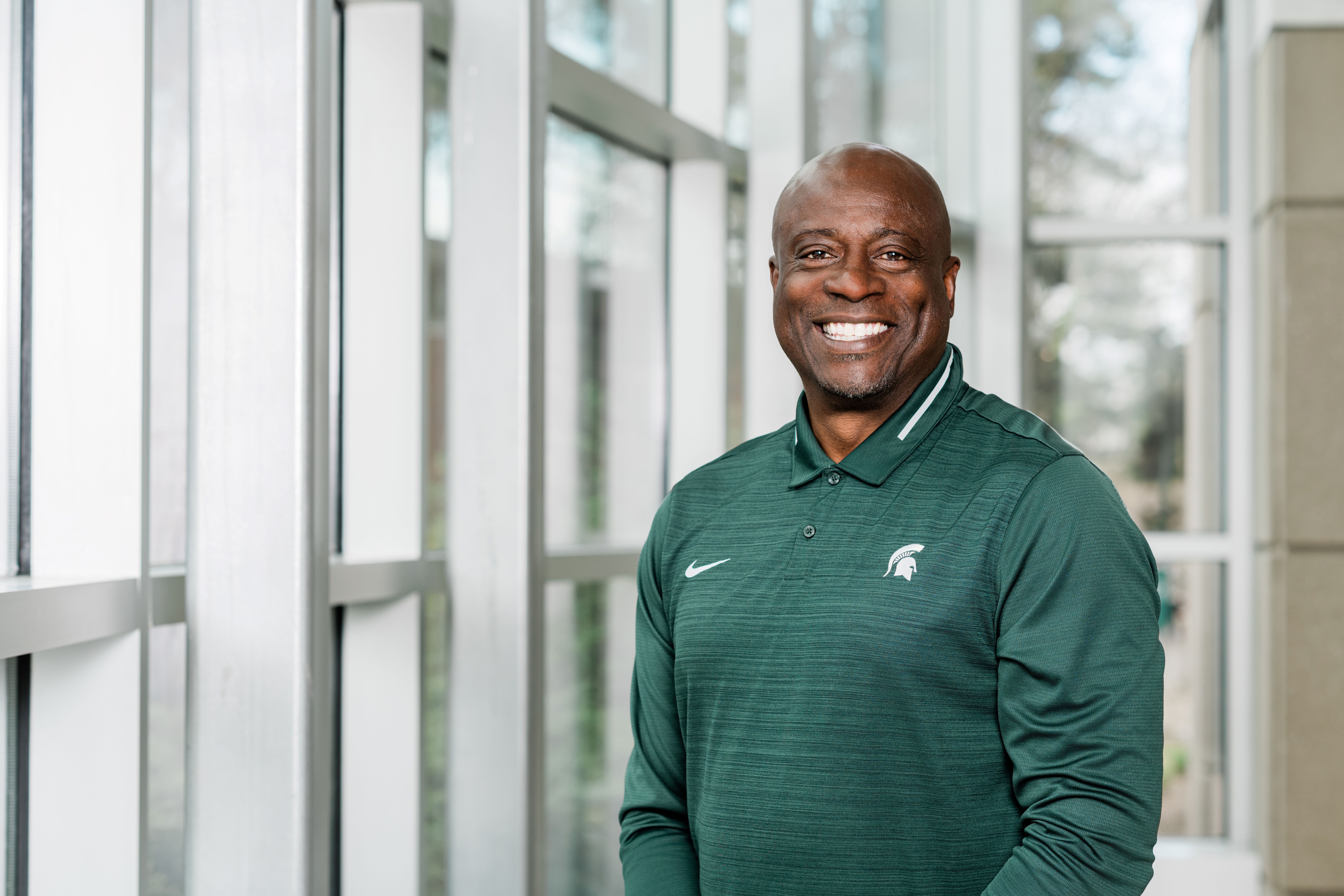 This image shows Sam Vincent, the 2024 Michigan State University Homecoming Grand Marshal, standing indoors near large windows. He is smiling broadly and wearing a green Michigan State Spartans polo shirt with a white Spartan helmet logo on the chest. The natural light from the windows brightens the space, and the background is softly blurred, keeping the focus on Sam Vincent. His confident and welcoming expression reflects the honor of his role as Grand Marshal.