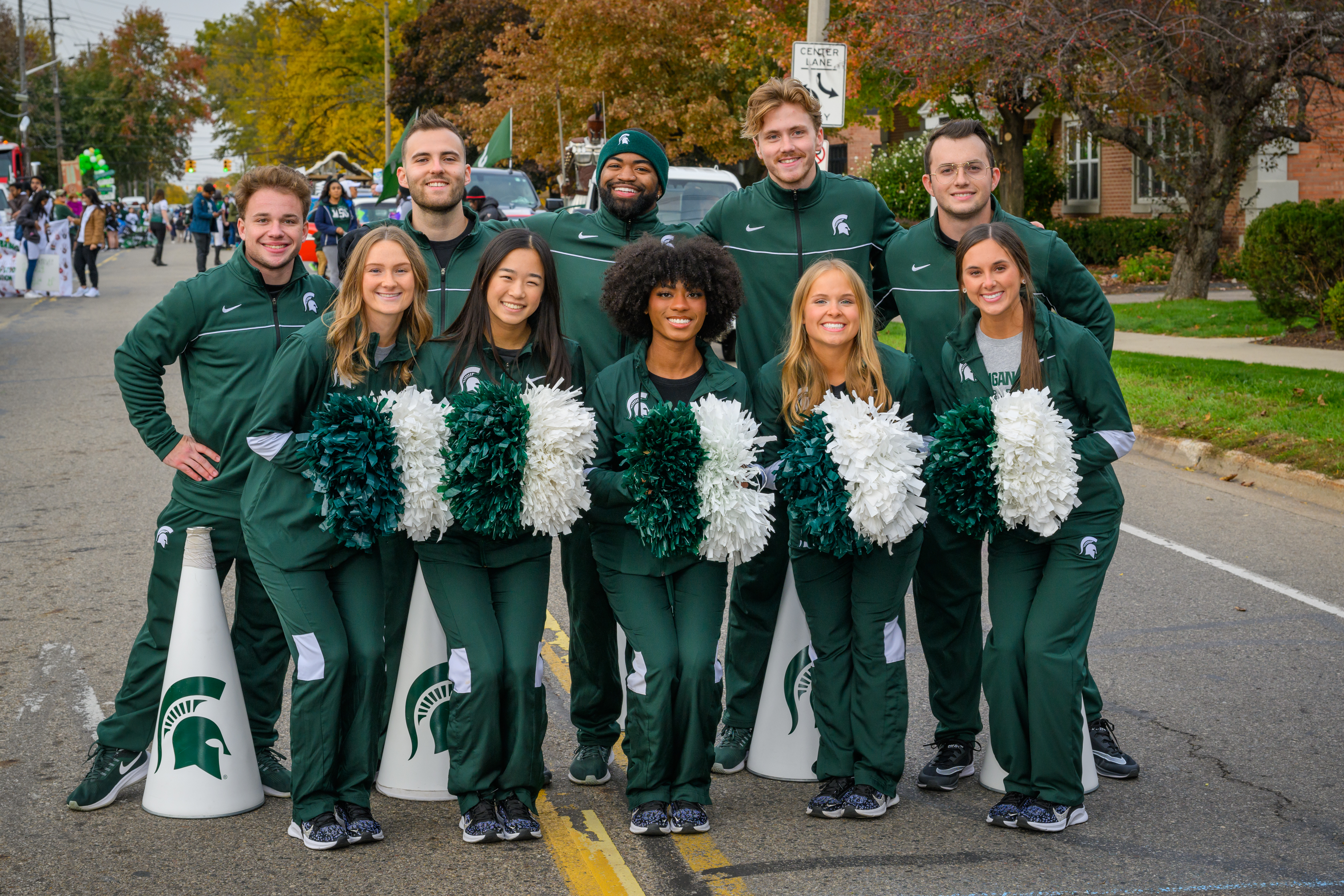 A group of nine Michigan State University cheerleaders, dressed in green tracksuits with the Spartan logo, pose together during the Michigan State University Homecoming Parade. The cheerleaders are holding white and green pom-poms and are standing in a row on a street lined with trees displaying fall foliage. In the background, there are other parade participants and spectators. The atmosphere is lively, with the cheerleaders smiling and showing team spirit.