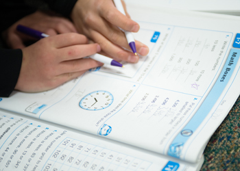 The hands of a child and an adult holding pens and working in a math book.