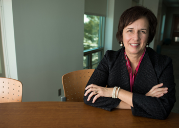 A women in business attire siting at a table with her arms folded, smiling..
