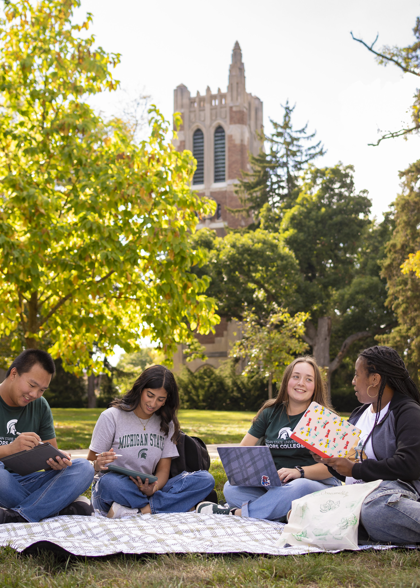 Photograph of Michigan State Students sitting on a blanket with books and computers on a sunny day. Beaumont Tower in the background.