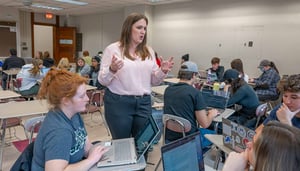 Students sit at desks with laptops in a classroom. An instructor stands and talks to them.