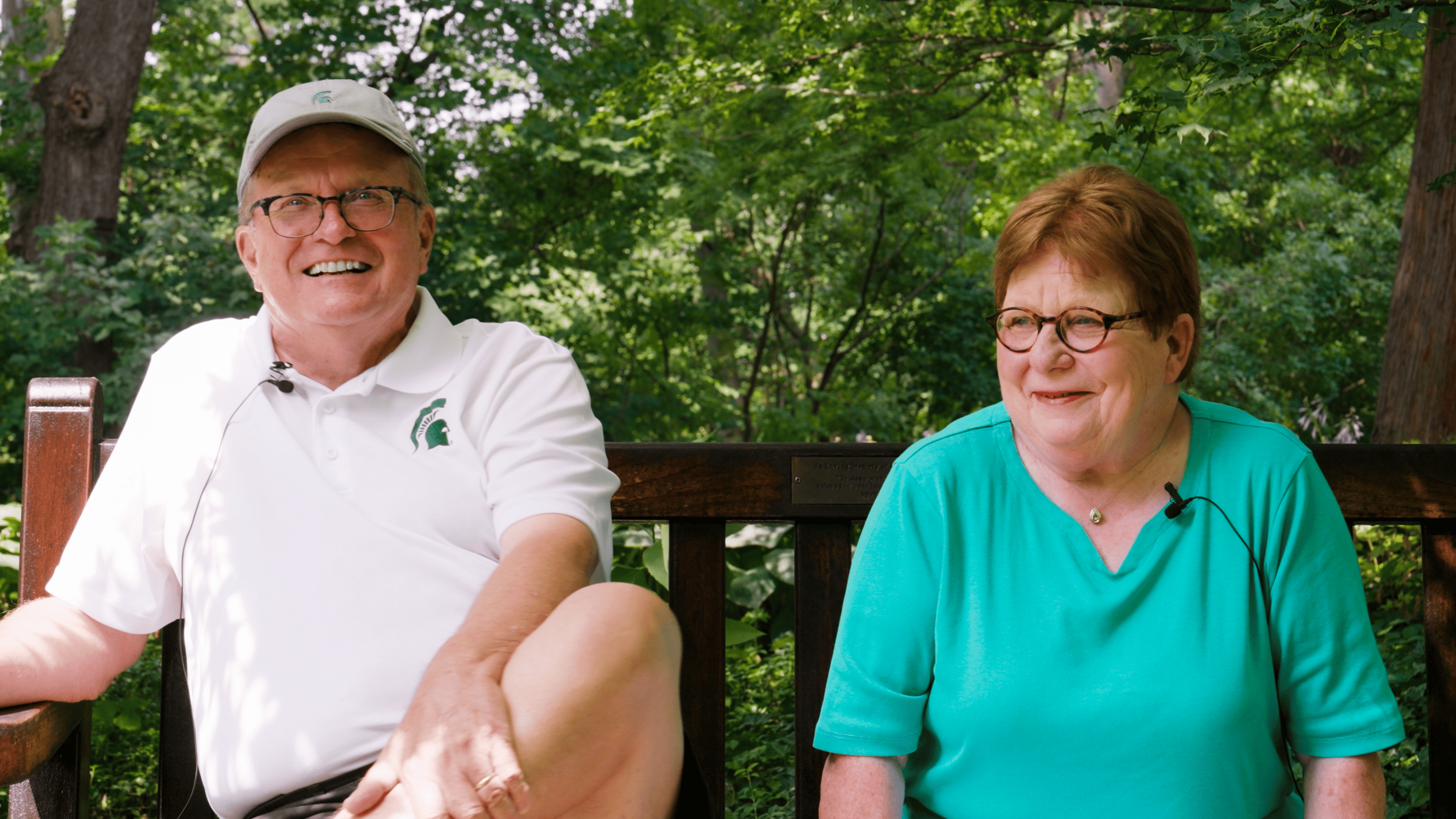 A man in Spartan shirt and woman in blue shirt sit on a bench in Beal Botanical Garden