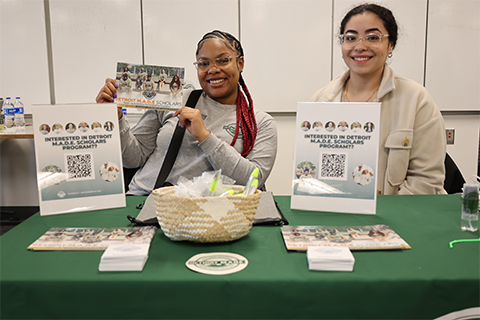 Two people sitting behind a table with table signs sitting on it, smiling and looking at the camera, while the person on the left holds up a postcard