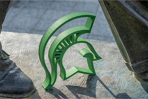 A green spartan helmet cutout sits at the feet of the John Hannah statue on MSU's campus