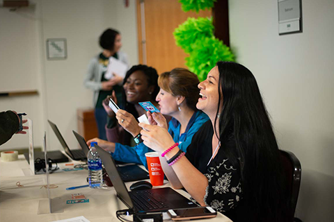 A row of students sitting in front of laptops smiling at each other and talking.