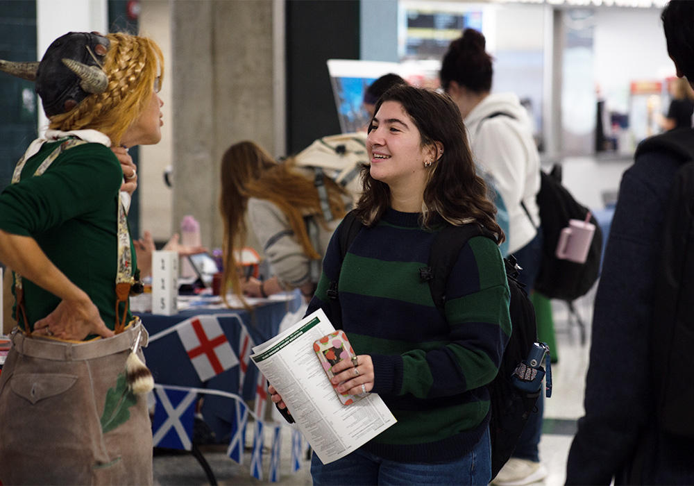 A student in a striped sweatshirt stands and talks with someone wearing a viking style helmet