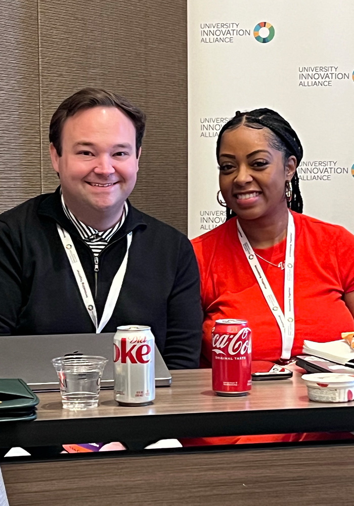 Two people sitting next to each other at a table with drinks and other items on it smiling at the camera