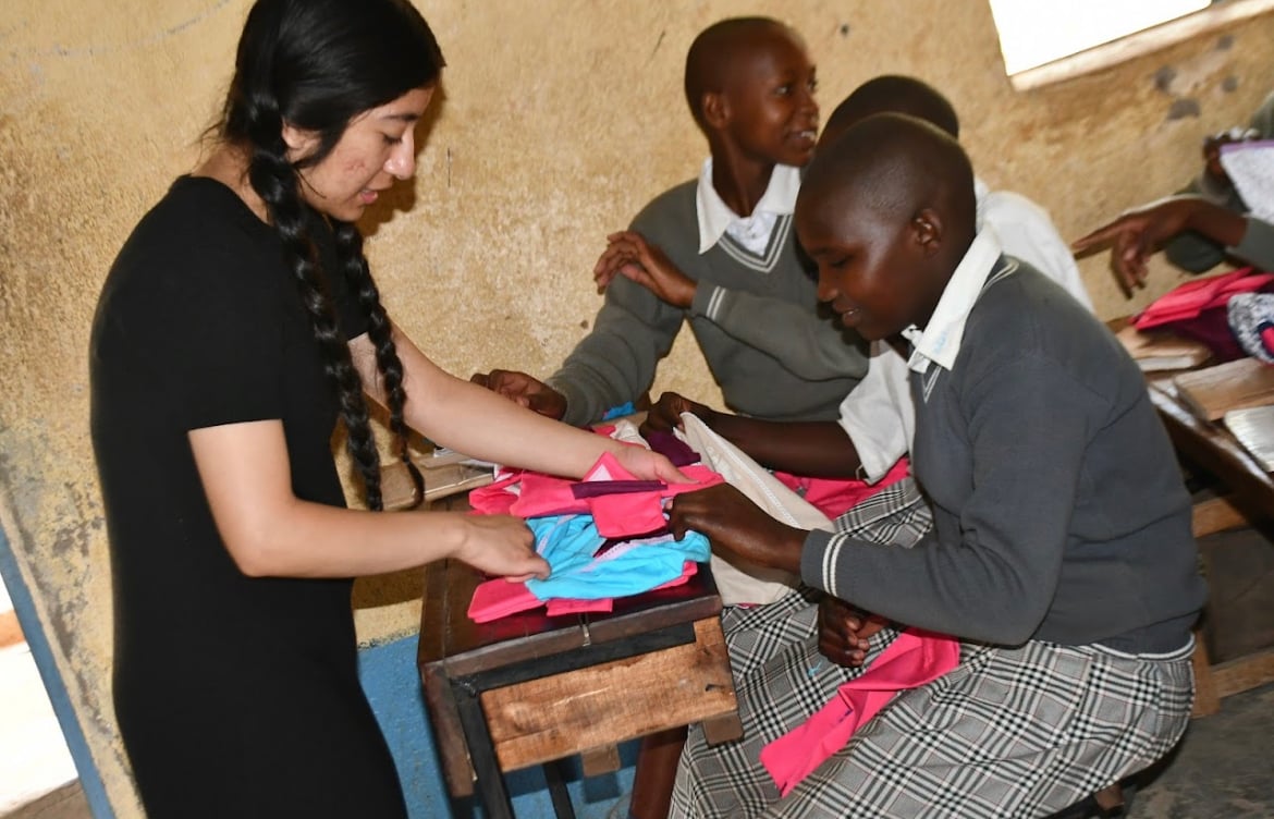 Faith standing in front of three students at at a desk explaining how to use the pink/red items they are holding.