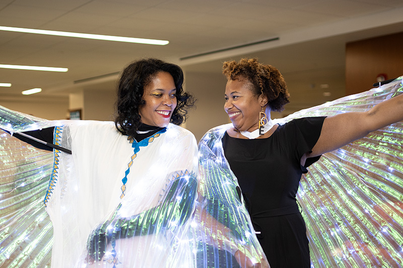 Two women stand side-by-side looking toward each other, each with one arm each lifted, while wearing sheer colored capes.