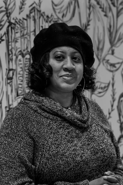 A black and white portrait of an individual looking just off center of camera. Their hair is chin length and curly and they are wearing a black floppy beret with a cowl neck sweater standing against a wall covered in drawings.