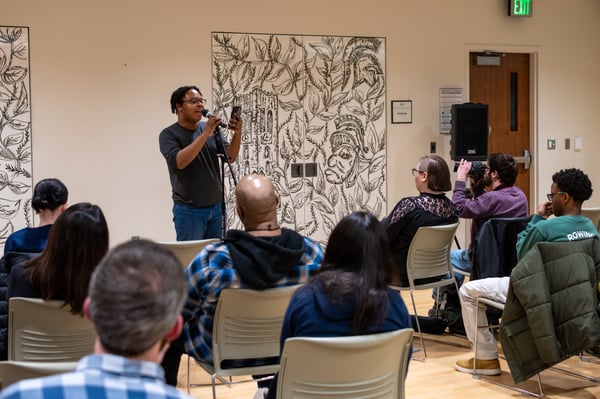 A person stands at a microphone with their arm outstretched in front of them holding a phone. They are in front of a small audience sitting in chairs. 