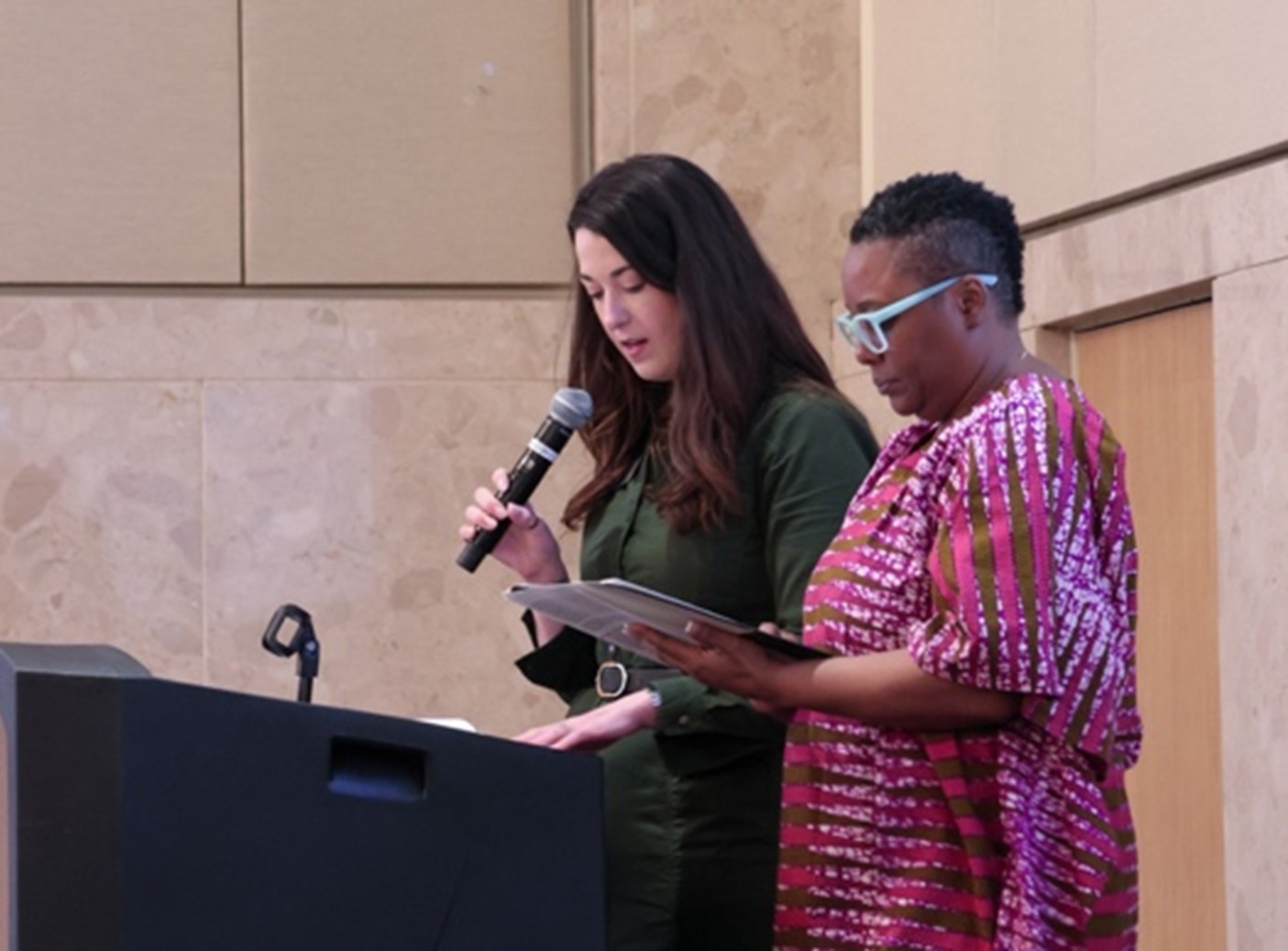 Dr. Qiana Green, wearing a pink and gold dress with light blue glasses, holds program documents while standing at the podium beside Katie Peterson who is wearing a belted dark green dress and speaking into a hand-held microphone.