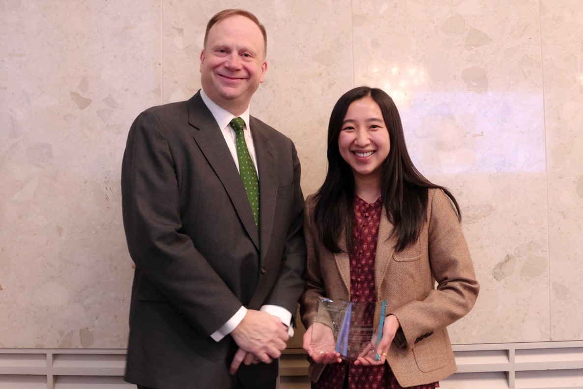 Heather Wilson, wearing a brown blazer over a red patterned dress shirt and holding a glass award plaque, stands smiling beside Mark Largent who is wearing a dark suit with a white dress shirt and green tie.