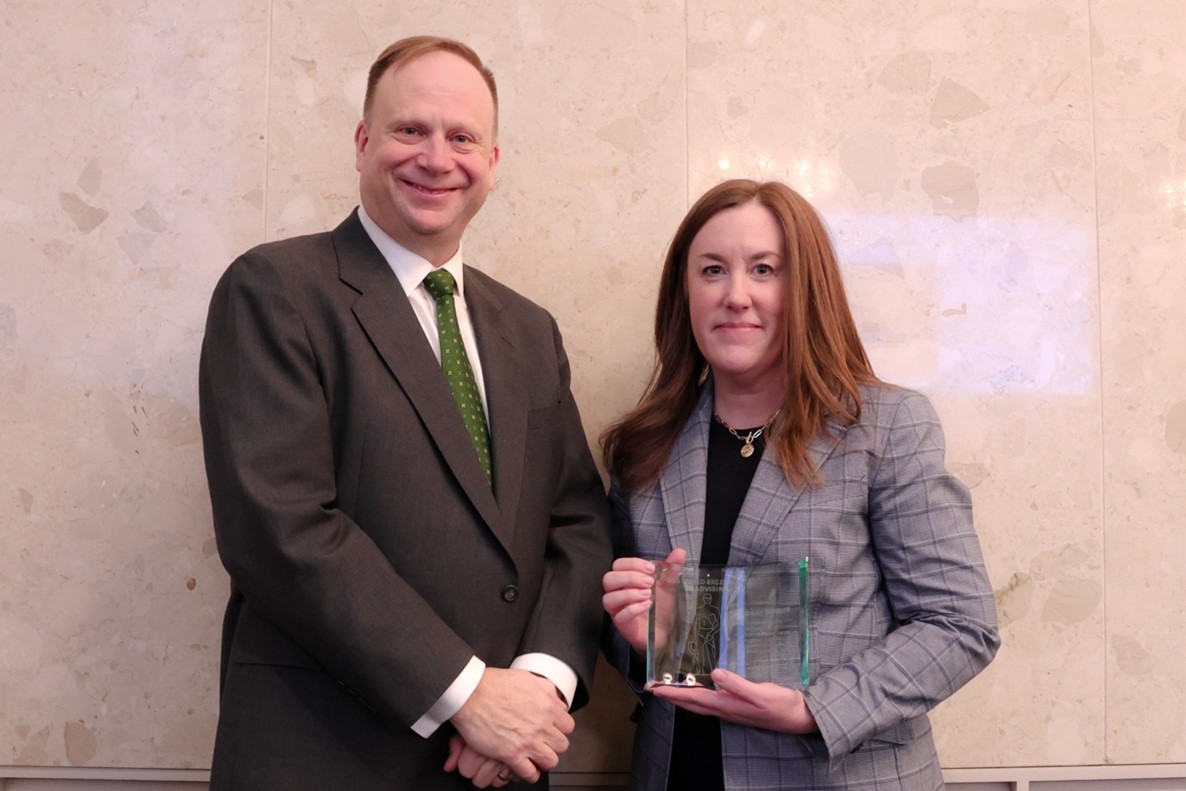 Lindsay Snyder, wearing a grey plaid blazer over a black shirt and holding a glass award plaque, stands beside Mark Largent who is wearing a dark suit with a white dress shirt and green tie.