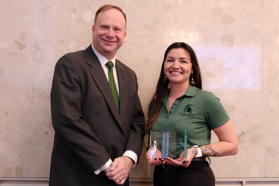 Erica Weeks, wearing a green MSU branded polo tucked into dark belted pants and holding a glass award plaque, stands smiling beside Mark Largent who is wearing a dark suit with a white dress shirt and green tie.