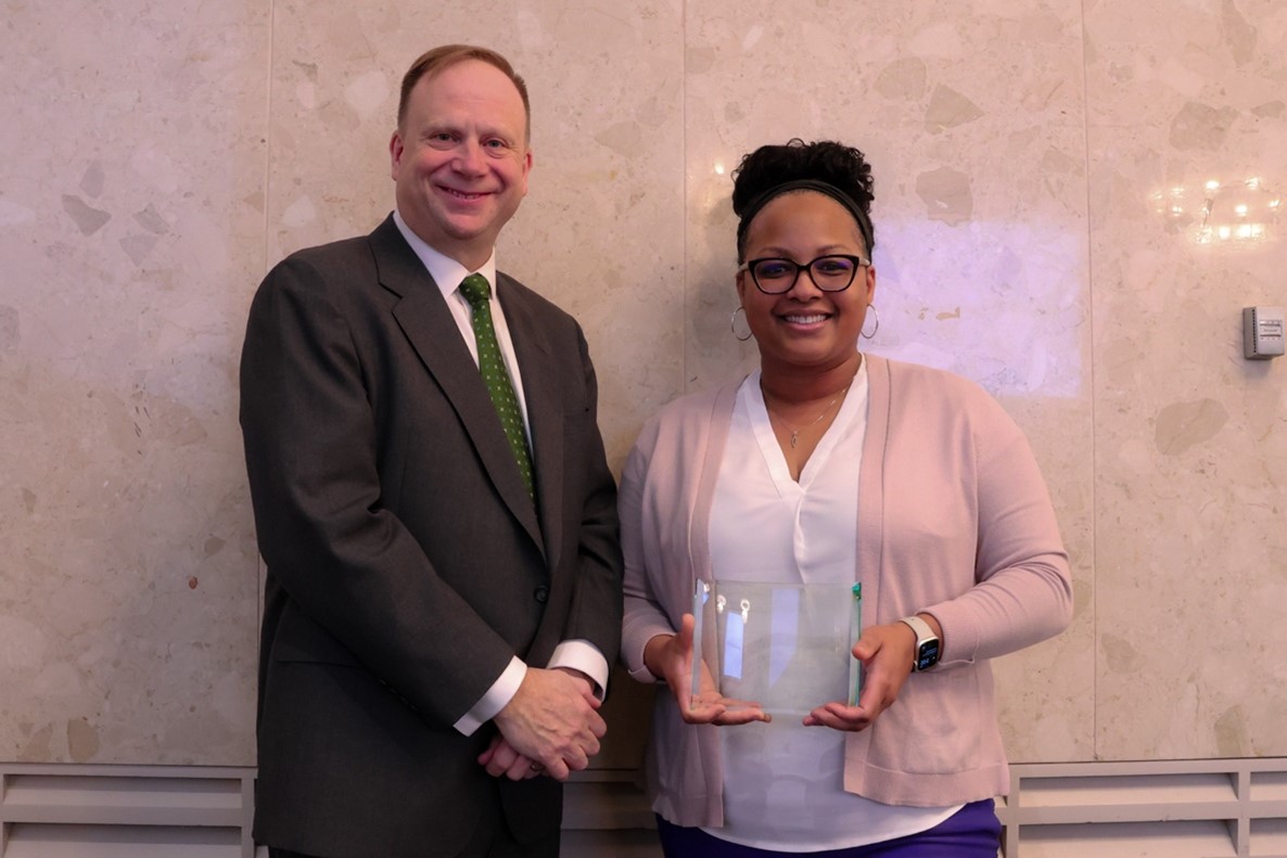Krystal Owens, wearing a pale pink cardigan over a white dress shirt and holding a glass award plaque, stands smiling beside Mark Largent who is wearing a dark suit with a white dress shirt and green tie.