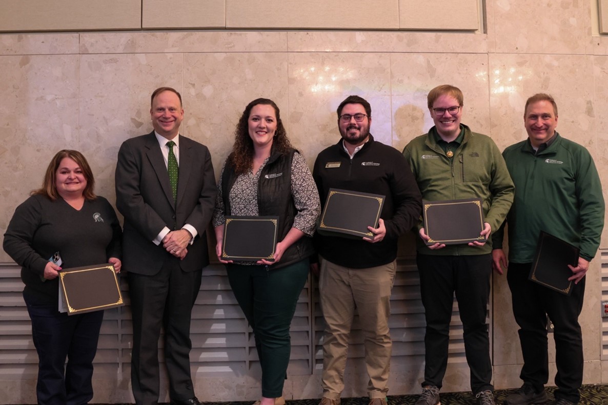 Representatives from the Institute of Agricultural Technology Program coordinators stand shoulder to shoulder, each holding their award certificate, with Mark Largent who is wearing a dark suit with a white dress shirt and green tie.