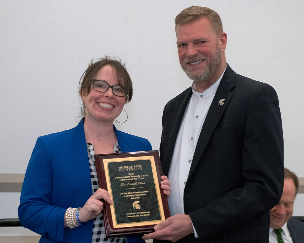 Interim Provost Thomas Jeitschko, wearing a white button-down shirt and a black jacket with a Michigan State University Spartan lapel pin, poses with Sarah Prior, wearing a blue blazer over a checkered shirt, both holding a square plaque honoring Prior as an Undergraduate Research Mentor of the Year winner.