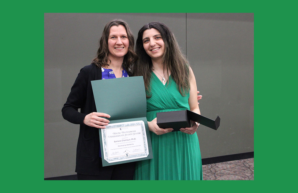 Two people standing next to one another, the person on the right with long dark hair and a green dress, and the person on the left wearing a dark sweater with short brown hair. They are both holding a certificate paper in their hands and displaying it to the camera while smiling.