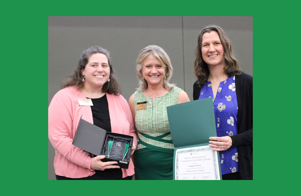 Three people standing together looking at the camera and smiling. The person on the right wearing pink is smiling and holding a small award in their hand. The person on the left, wearing black with a purple shirt is holding and displaying a certificate.