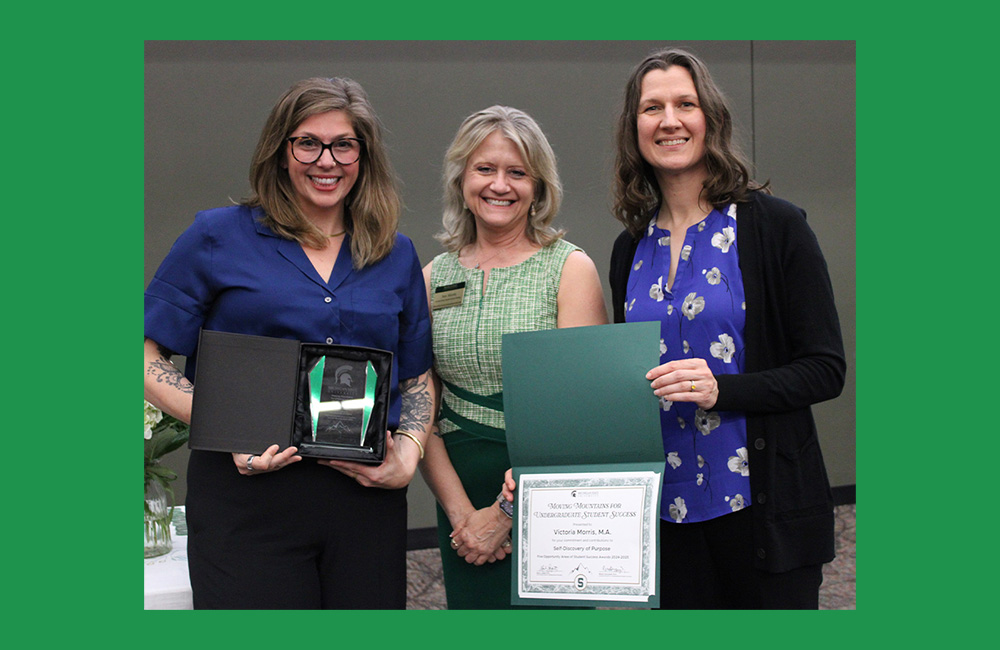Three people standing together smiling at the camera. The person on the left, wearing dark purple, is holding a small square award. The person on the right, wearing purple and black is holding a paper certificate in a green folder.