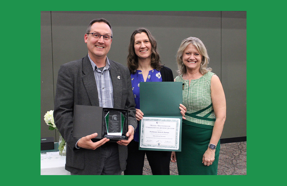 Three people standing next to one another smiling at the camera. The person on the left is wearing a grey suit jacket holding a small square award. The person in the center, wearing purple and black, is holding a paper certificate in a green folder. 