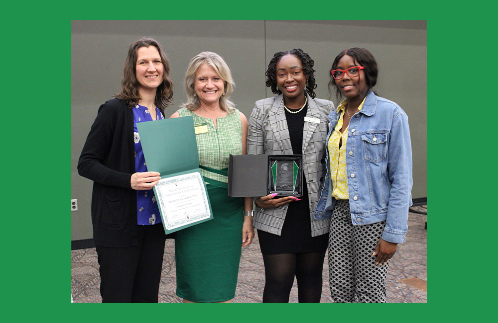 Four people standing next to one another smiling at the camera. The person at the end is holding a paper certificate in a green folder. The person second to the right is holding a small square award.