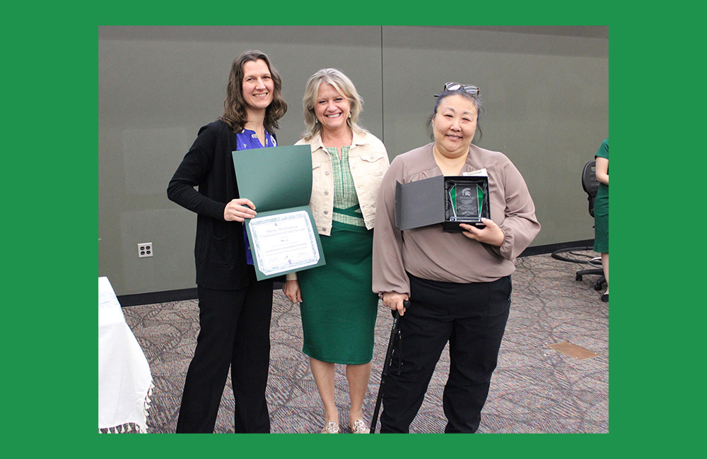 Three people standing next to one another smiling at the camera. The person on the left, wearing black and purple, is holding a paper certificate in a green folder. The person on the end is holding a small square award.