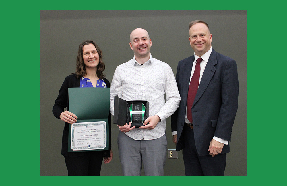 Three people stand next to one another smiling at the camera. The person on the left, wearing black and purple, is holding a paper certificate in a green folder. The person in the center, wearing a white shirt, is holding a small square award.