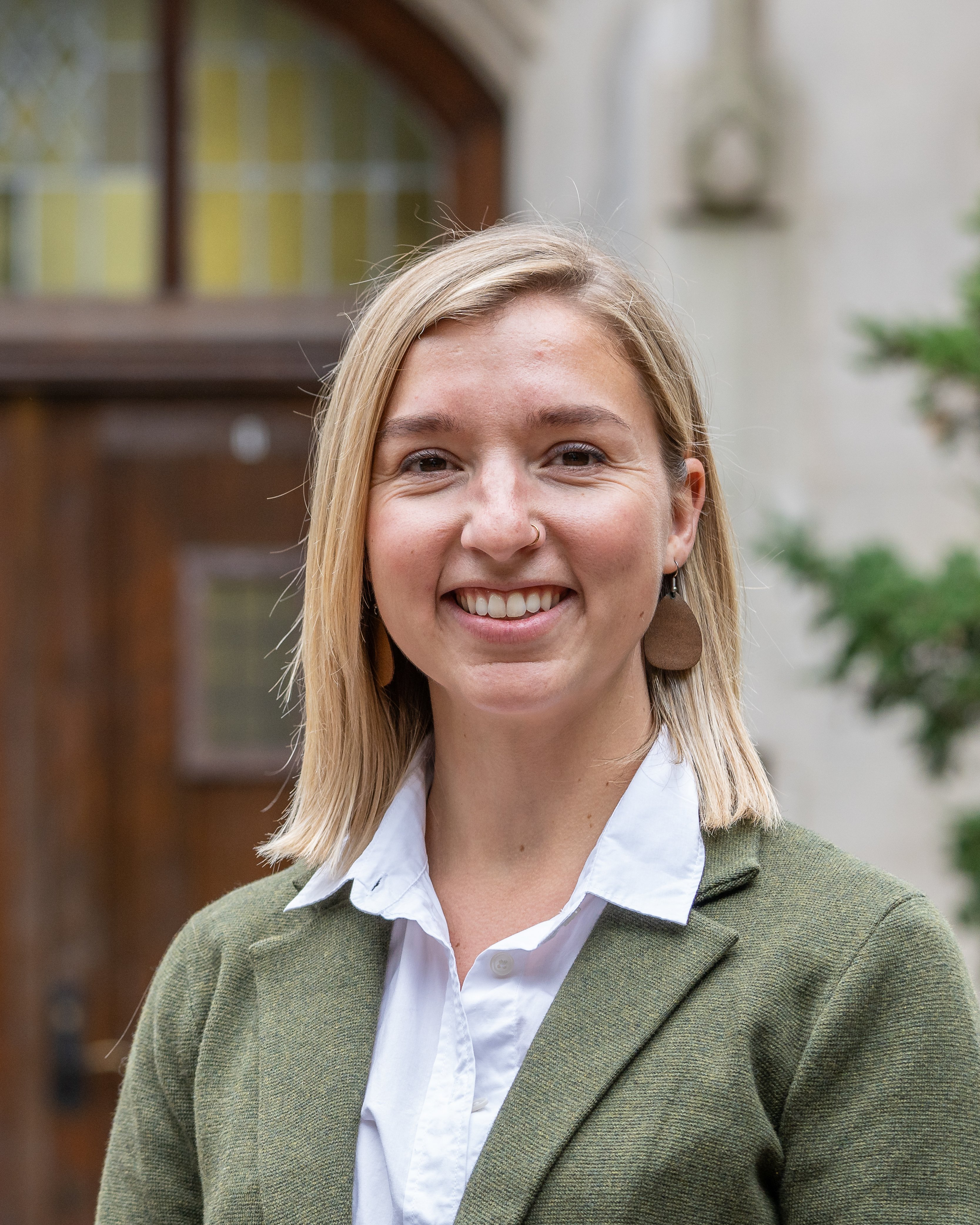 Portrait photo. The subject is looking at the camera smiling. They have straight blonde hair shoulder length and are wearing a green cardigan over a white collared shirt.