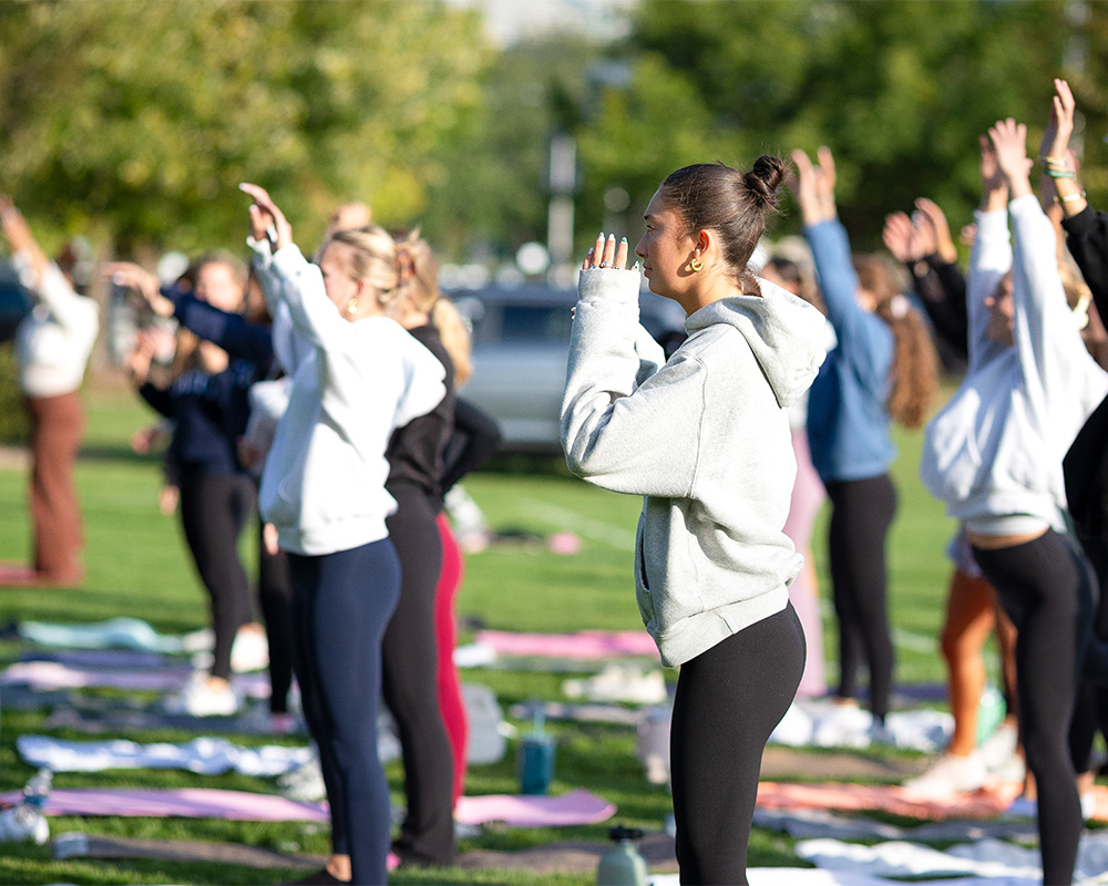 A group of people stand in rows on yoga mats. Most are wearing sweatshirts, and their arms are stretched above their heads toward the sky. They are standing in a grassy field on a sunny day.