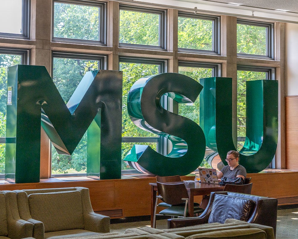 Large green block letters spelling "MSU" are placed in front of tall windows in the MSU Union. A student sits in front of the letters at a small table showcasing how tall they are.