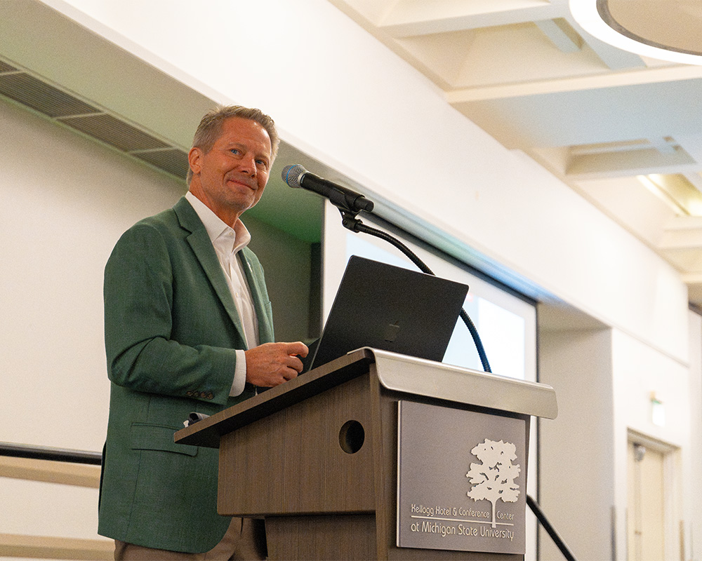 MSU President Guskiewicz stands behind a podium, with a microphone pointed toward him and a computer open in front of him. He is looking off into the distance beyond the camera. He is wearing a grey suit jacket and a white shirt.