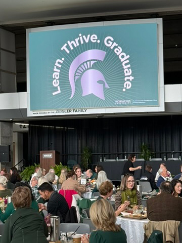 Large presentation screen above people sitting at circle tables. The screen has a large Spartan helmet on it and has the words Learn, Thrive, Graduate in large feature above the helmet.