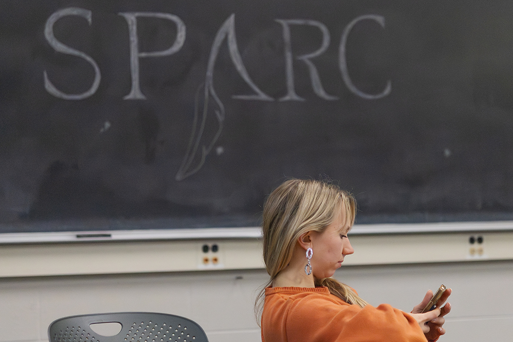A student sits to the right of the frame facing right, looking down at their phone in their hands, while wearing a an orange sweater with blonde hair swept over their left shoulder. They are sitting in front of a large black chalkboard with the word SPARC written in white chalk.