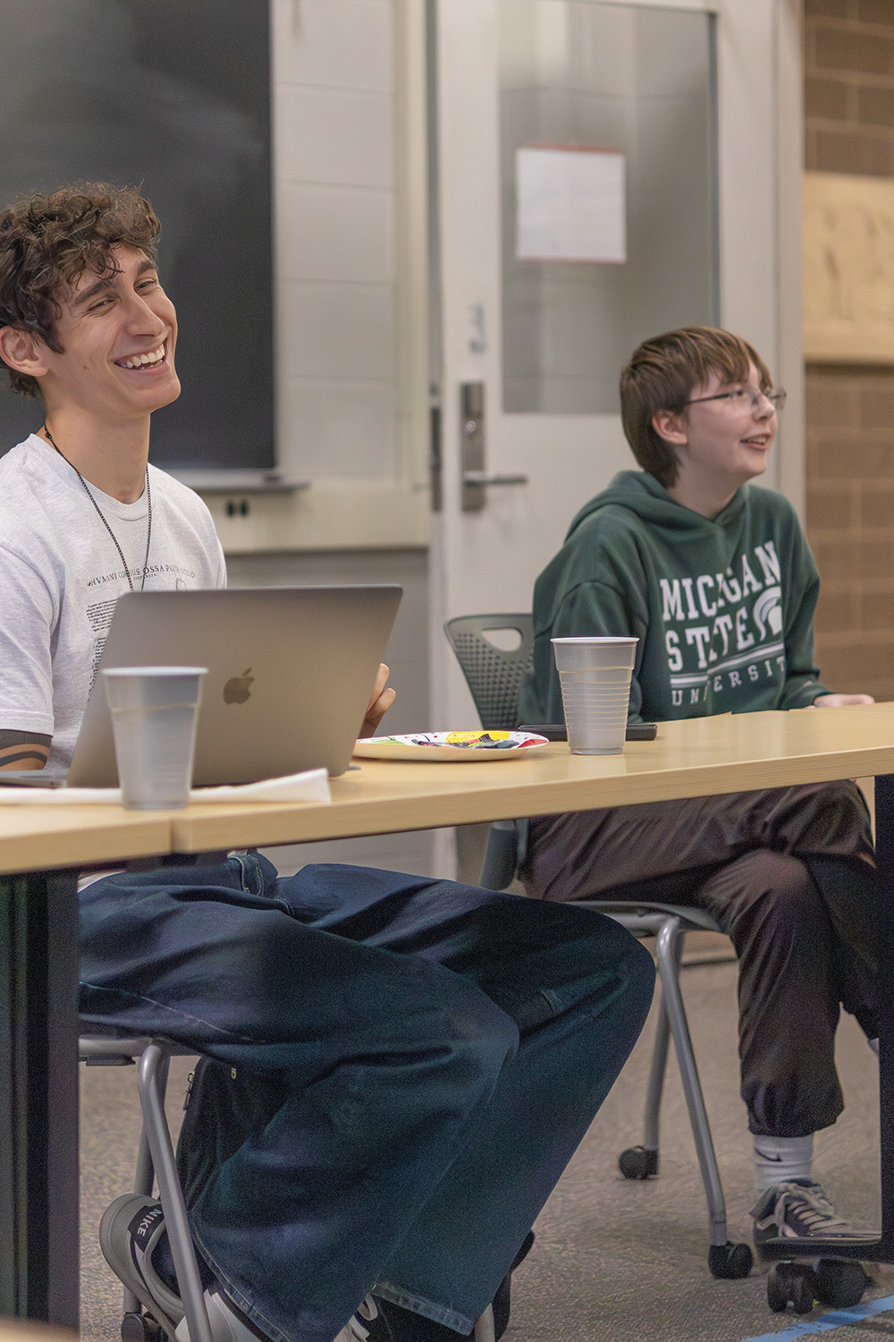 Two students sit at a wooden table looking across at something out of frame. The student on the right is smiling broadly, and sits in front of an open computer. The student on the right also smiles and is wearing a green shirt.