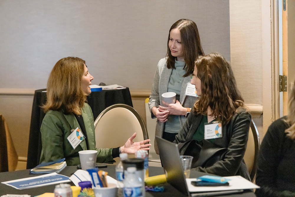 Three people are having a conversation. Two are sitting at a large banquet table looking up at another person who is standing and holding a to-go coffee cup.