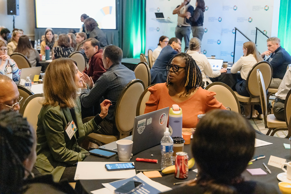 View of a busy room with circle banquet tables in black table cloths covered in papers, pens, computers and notebooks. The individuals are talking and facing each other. The image feature twos two individuals speaking to one another at the center.