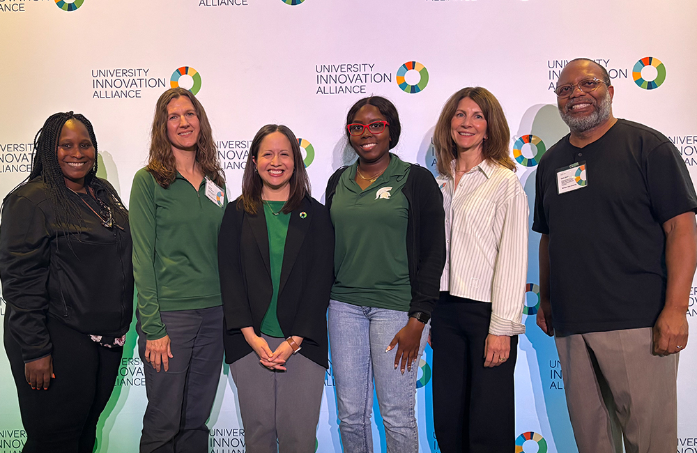 Six people stand shoulder to shoulder while looking at the camera and smiling. They are standing in front of a UIA backdrop.