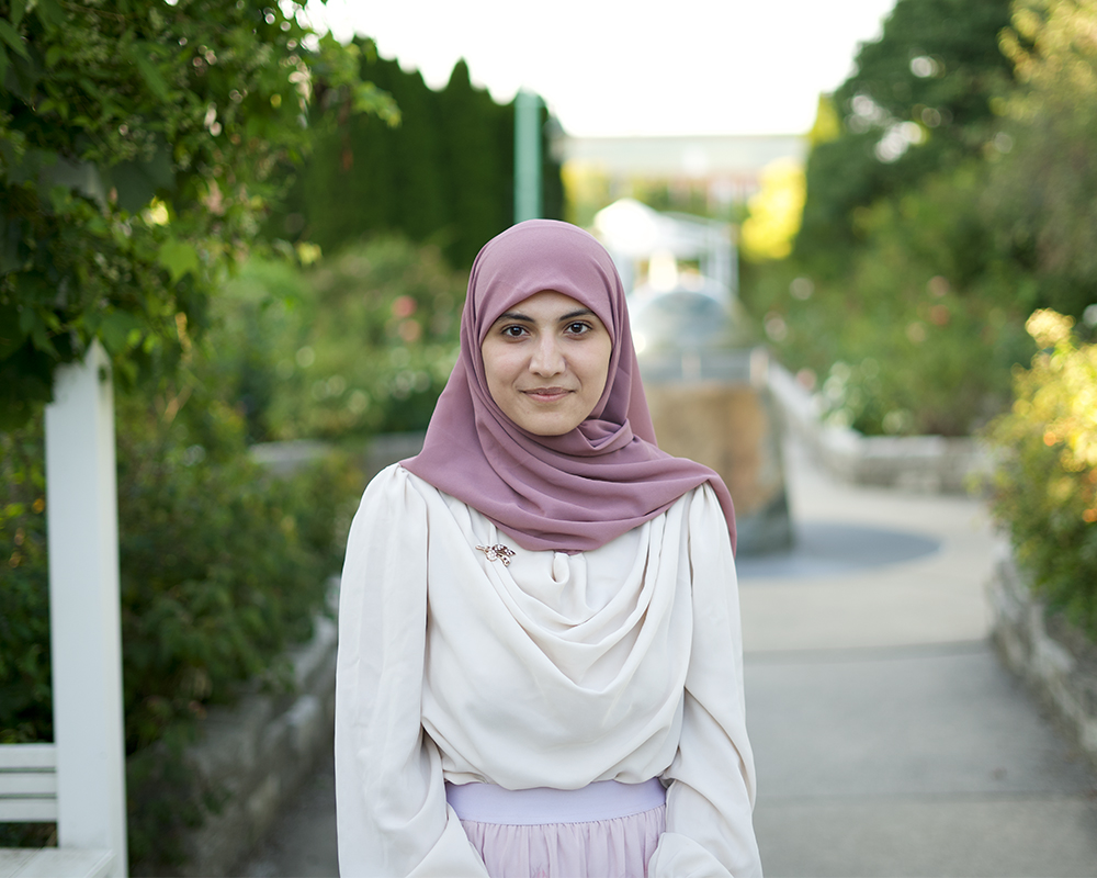 Portrait. Subject is standing on a walkway with green foliage on either side. They are looking directly at the camera with a small smile, wearing a white, long-sleeved, draped shirt with a mauve scarf wrapped around their head.