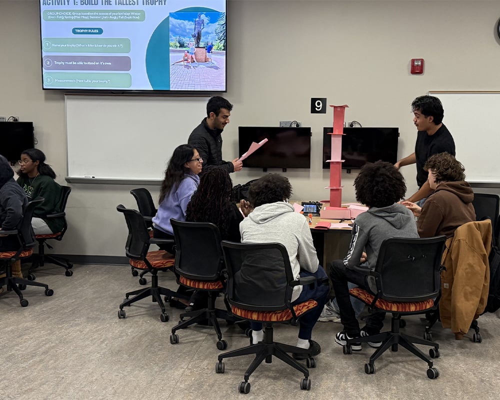 Students sit in roller chairs around a table while two individuals stand and lean in toward the middle at a tall paper object. They are in a classroom setting with a large digital screen displaying in the background.