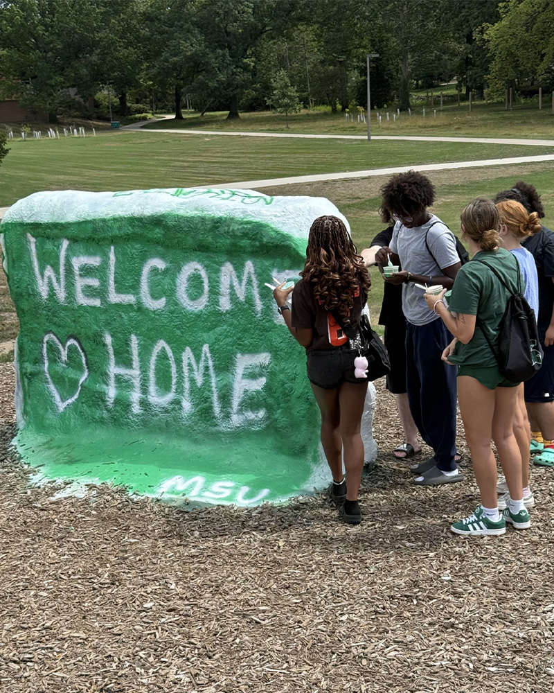 A small group of students stand to the right of MSU's rock which is painted green with white words reading 