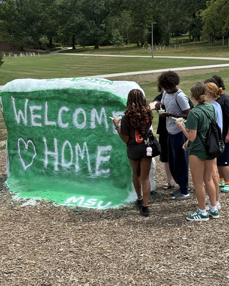 A small group of students stand to the right of MSU's rock which is painted green with white words reading "Welcome Home" with a heart shape.
