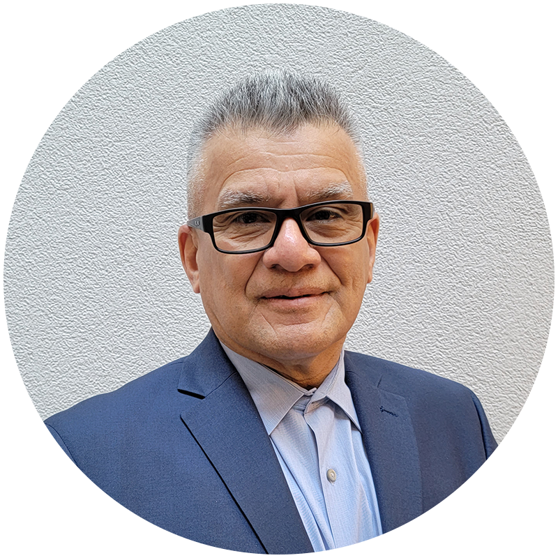 Portrait image in a circular frame. Subject is looking at the camera and smiling, seen from the chest up. They are wearing a blue suit jacket and light blue collared shirt with dark framed glasses and short, grey hair.