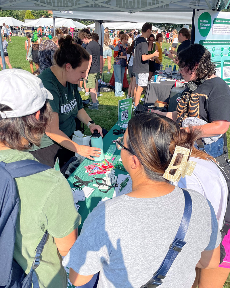 Students gather around a person at a table looking through items in front of them. There is a sign on the table reading Spartan Experience Record with a QR code to scan. 