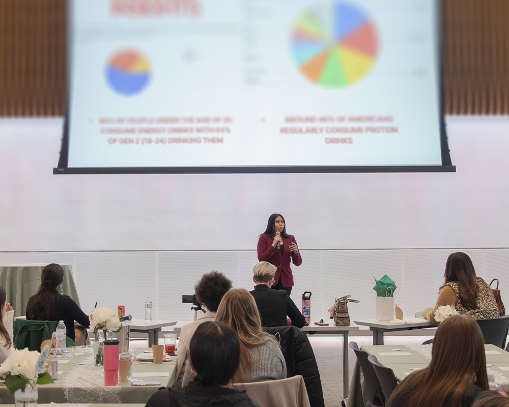 An individual stands in front of a large presentation screen speaking directly to individuals sitting a banquet tables looking at the presentation. 