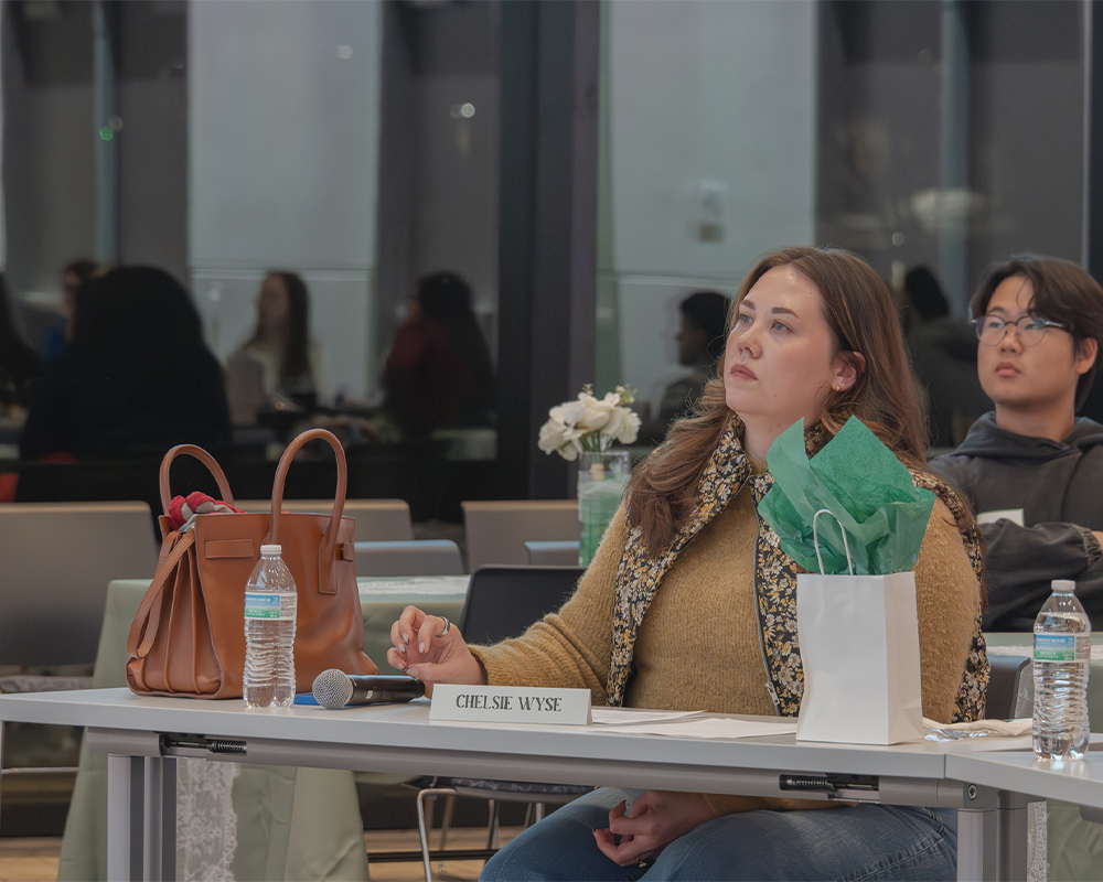 An indivdiviaul sits at a table looking somewhere out of frame. They are holding a pen in hand, sitting at a table with a name plate in front of them, wearing a tan shirt and vest.