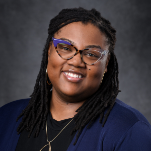 Portrait. Subject is seen from the shoulders up. They are wearing a blue sweater with a black shirt, smiling and looking directly at the camera. Long braided hair is coming down their shoulders. 