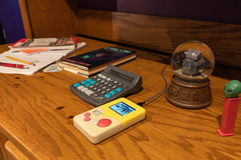 Various items, including what looks like a calculator, Game Boy, and snow globe and Pez dispenser sit on a wooden desk with notebooks, paper and pencils scattered around.